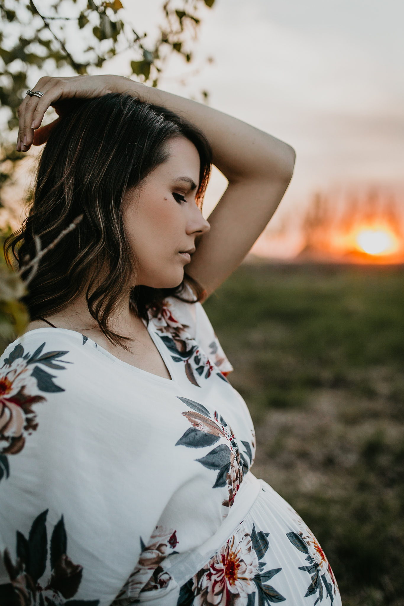 Expecting woman by a tree with one arm up over her head in front a fire orange sky.